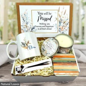 May include: A gift box containing a white ceramic mug with the name Olivia and a floral design, a Peaceful Zen soy wax candle in a metal tin, a silver-toned spoon with a floral handle, and a square coaster with a multi-colored wood grain pattern.