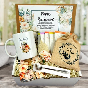 May include: A retirement gift box with a white mug personalized with "Michelle" and floral design. Includes tea bags, a burlap bag with "Tea" in a wreath, a spoon, and a card that reads "Happy Retirement". The box is filled with decorative flowers.