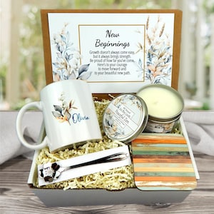 May include: A gift box with a white mug, a candle, a coaster, and a spoon. The mug has a floral design and the name "Olivia". The candle is in a metal tin. The box includes a card with the text "New Beginnings".