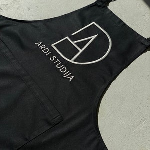 Può includere: Grembiule nero con un logo ricamato bianco che dice "ARDI STUDIJA" e un design geometrico.