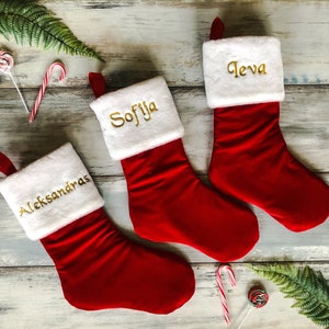May include: Three red Christmas stockings with white cuffs and gold embroidered names: Aleksandra, Sofija, and Ieva.
