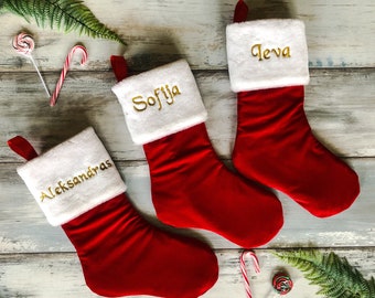 May include: Three red Christmas stockings with white cuffs and gold embroidered names: Aleksandra, Sofija, and Ieva.