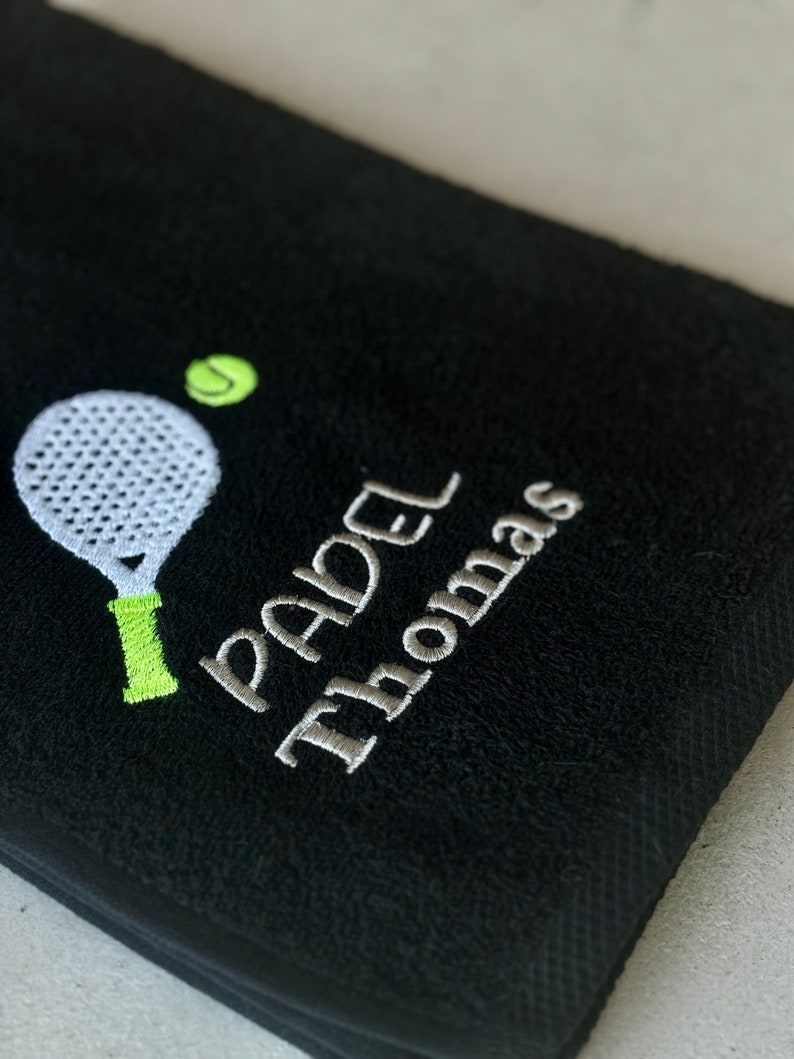 Puede incluir: Toalla negra con bordado blanco de una raqueta de p&aacute;del y una pelota, y el texto "PADEL THOMAS".