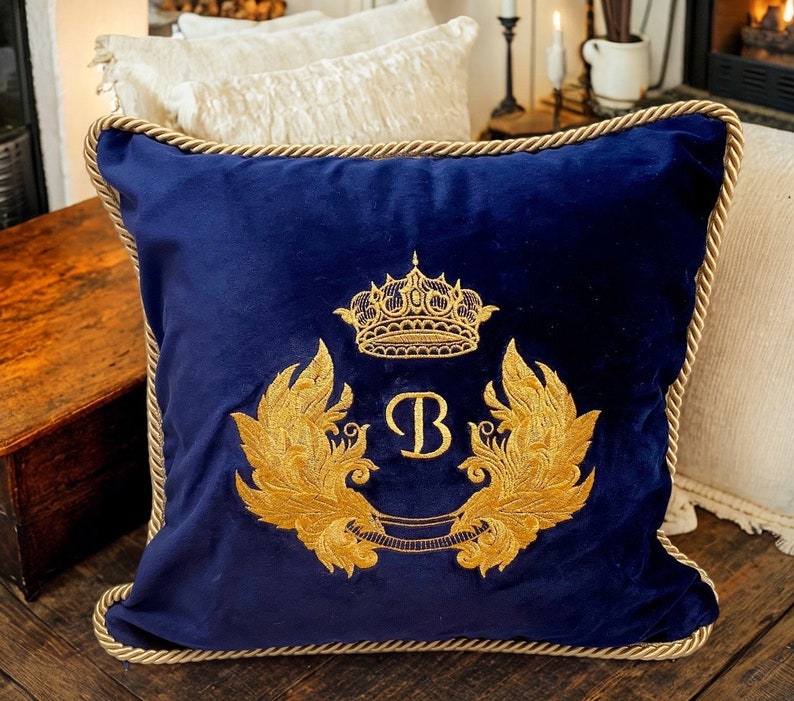 May include: A navy blue velvet pillow with a gold embroidered crown, initial "B", and ornate wings. The pillow has a gold rope trim.