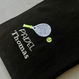 Puede incluir: Toalla negra con un dise&ntilde;o de pala y pelota bordados en blanco y verde. El texto "PADEL Thomas" est&aacute; bordado en verde.