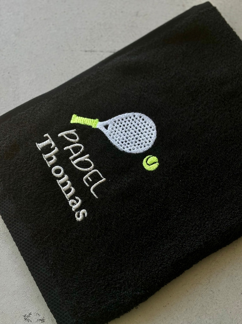 Puede incluir: Toalla negra con un dise&ntilde;o de pala y pelota bordados en blanco y verde. El texto "PADEL Thomas" est&aacute; bordado en verde.