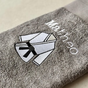 Könnte beinhalten: Graues Handtuch mit gesticktem Karate-Anzug-Design in Weiß und Schwarz. Der Name "Matheo" ist in weißer Schrift gestickt. Das Handtuch ist gefaltet und hat eine strukturierte Optik.