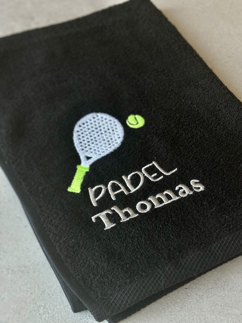 Puede incluir: Toalla negra con un paddle y una pelota bordados en blanco y verde, y el texto "PADEL Thomas" en blanco.