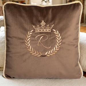 Può includere: Un cuscino in velluto marrone con un design a corona e corona d'alloro ricamata in oro. La corona circonda la lettera "R". Il cuscino ha un bordo in corda dorata.