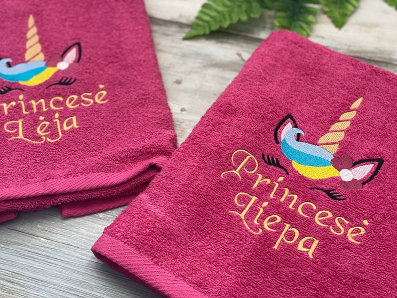 May include: Two pink towels with embroidered unicorn designs and personalized names. The towels feature a gold unicorn with a rainbow mane and a flower on its horn. The names "Princes&eacute; Leja" and "Princes&eacute; Liepa" are embroidered in gold.