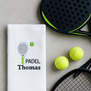Può includere: Asciugamano da padel bianco ricamato con un disegno di racchetta e pallina da padel, la parola "PADEL" e il nome "Thomas" in nero. Sono visibili anche due racchette da padel nere e due palline da tennis gialle.