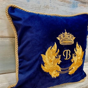 May include: A navy blue velvet pillow with a gold embroidered design. The design features a crown, a letter "B", and two stylized wings.