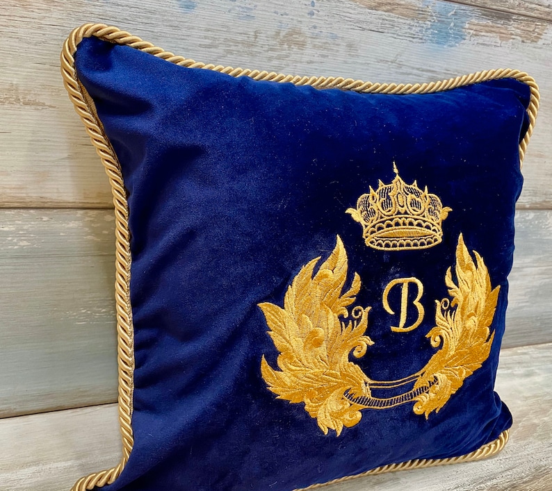May include: A navy blue velvet pillow with a gold embroidered design. The design features a crown, a letter "B", and two stylized wings.