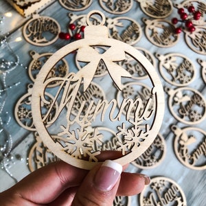 Può includere: Un ornamento natalizio in legno con la parola "Mamma" scritta su di esso. L'ornamento è decorato con fiocchi di neve e un fiocco.