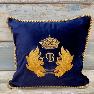 May include: A navy blue velvet pillow with a gold embroidered crown, wings, and the letter "B".