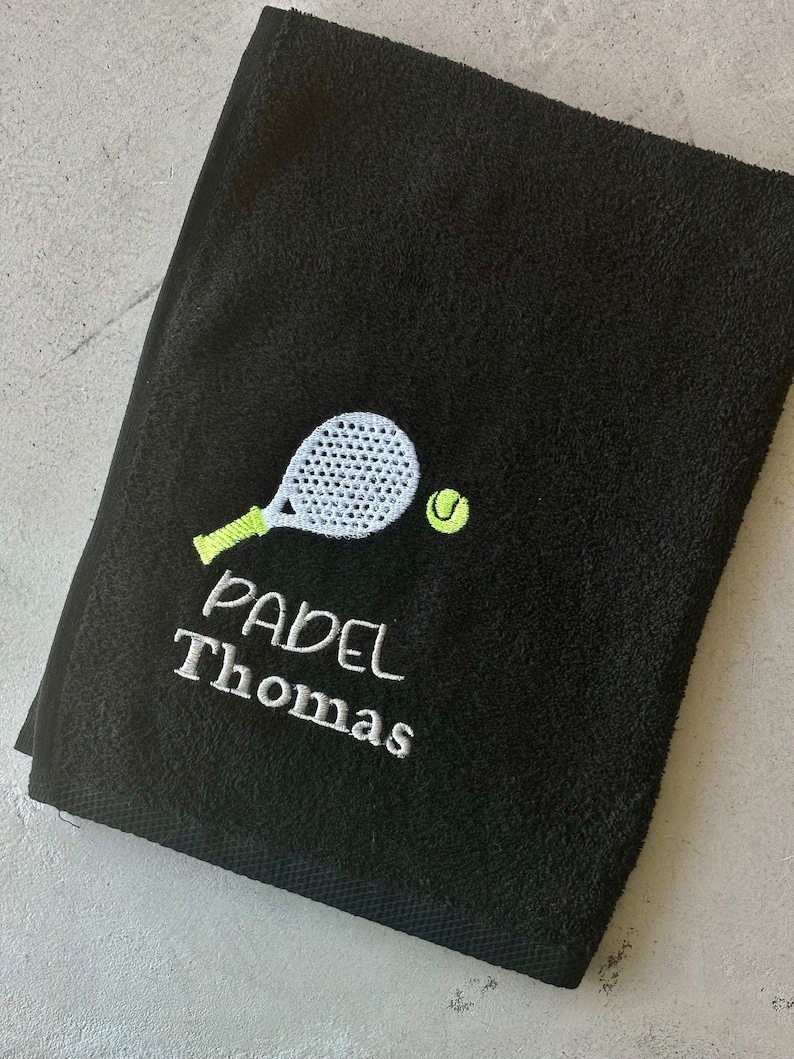 Puede incluir: Toalla negra con bordado blanco de una raqueta de p&aacute;del y una pelota. El texto "PADEL Thomas" est&aacute; bordado debajo de la raqueta.