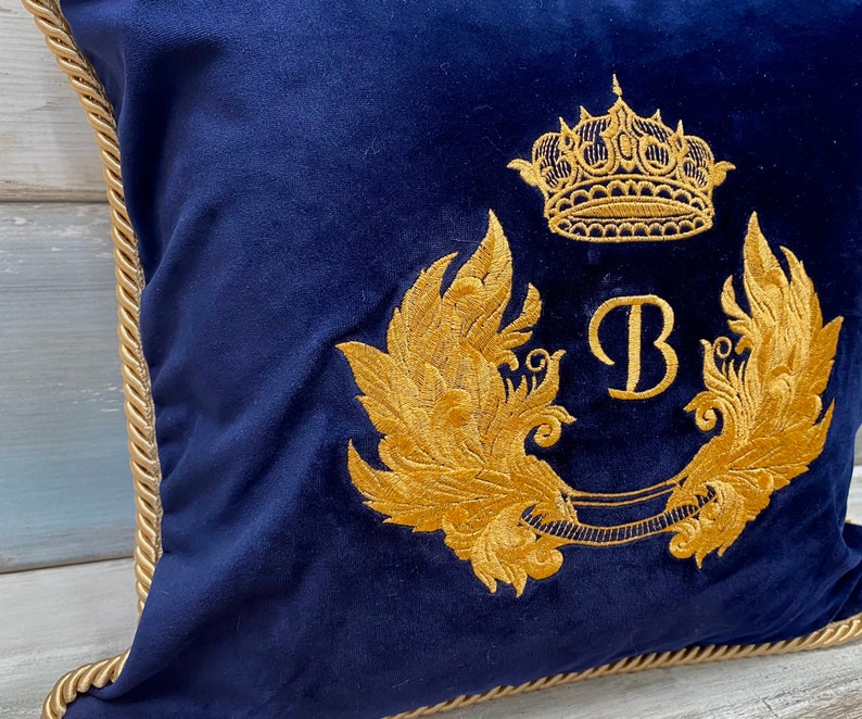 May include: A navy blue velvet pillow with a gold embroidered crown, wings, and the letter "B".