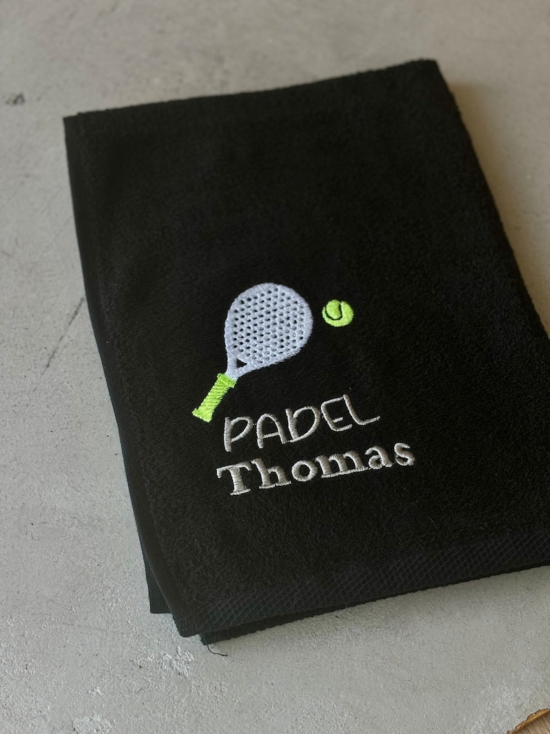 Puede incluir: Toalla negra con una pala bordada en blanco y una pelota de tenis verde. El texto "PADEL Thomas" est&aacute; bordado en blanco debajo de la pala.