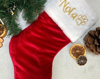 May include: Red velvet Christmas stocking with a white faux fur cuff. The cuff has the name "Nola" embroidered in gold thread with a paw print.