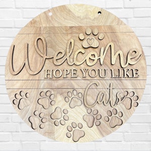 Könnte beinhalten: Runder Holzschild mit der Aufschrift "Welcome, Hope You Like Cats" und Pfotenabdrücken. Das Schild besteht aus hellem Holz mit natürlicher Maserung. Ein Herz befindet sich in der Mitte des Pfotenabdrucks.