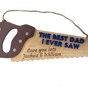 Könnte beinhalten: Sägeförmiges Holzschild mit braunem Griff und heller Holzblatt. Der Text "THE BEST DAD I EVER SAW" ist blau, darunter "Love you lots Joshua & William". Eine Juteschnur ist zum Aufhängen befestigt.