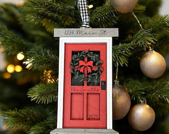 May include: A red door Christmas ornament with a wreath and bow, the text "The Smiths" and "2025". The ornament also has the text "1234 Main St" and "Our New Home". The ornament is hanging on a Christmas tree.