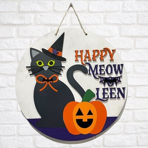May include: A round wooden door hanger with a black cat wearing a witch hat and holding a pumpkin. The text "Happy Meow-Leen" is written in black and orange letters.