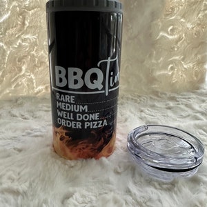 May include: Black and orange stainless steel tumbler with a gray lid and a white graphic that says "BBQ Time" with the text "Rare, Medium, Well Done, Order Pizza" underneath. The tumbler is sitting on a white fuzzy surface with a clear lid next to it.