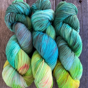 Peut inclure: Trois écheveaux de fil teints à la main dans des tons de vert, de bleu et de jaune. Le fil est disposé sur une surface en bois, avec les couleurs se fondant et créant un effet de dégradé. Le fil semble doux et texturé, adapté aux projets de tricot ou de crochet.