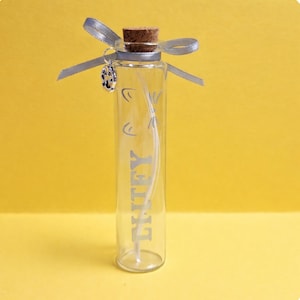 May include: A clear glass bottle with a cork stopper and a silver ribbon bow. The bottle has the word "LIFEY" printed vertically in silver. A small silver charm hangs from the ribbon. The background is a solid yellow.
