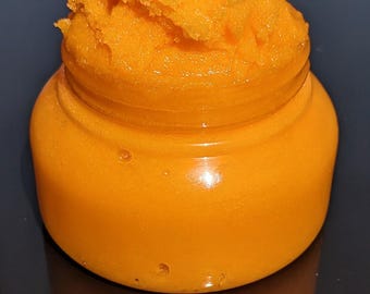 May include: A small, round, clear glass jar filled with a bright orange, textured substance. The jar is reflected in the dark surface below. The substance appears to be a cosmetic product, possibly a scrub or balm.