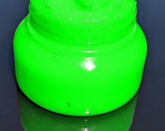 May include: A bright green jar filled with a textured, lime-green substance. The jar is round with a screw-top lid. The product and its reflection are visible against a dark background. The jar's glossy surface reflects light.