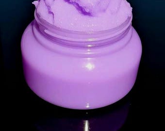 May include: A jar of lavender-colored body scrub. The scrub is a light purple color and has a textured, granular appearance. The jar is clear plastic, and the product is reflected on the black surface below.