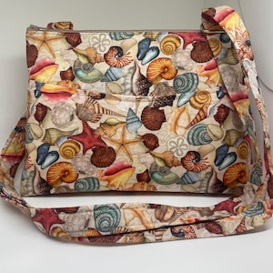 May include: A beige fabric crossbody bag with a long adjustable strap. The bag is decorated with a colorful print of seashells and starfish.