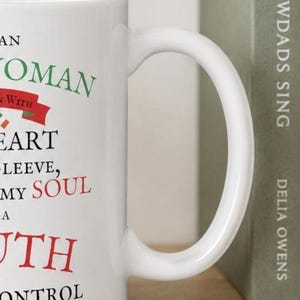 May include: A white ceramic mug featuring black, green, and red text that reads: AN WOMAN WITH HEART SLEEVE, MY SOUL A UTH ONTROL. The mug has a curved handle and a smooth finish, displaying a portion of the printed message on its side surface.