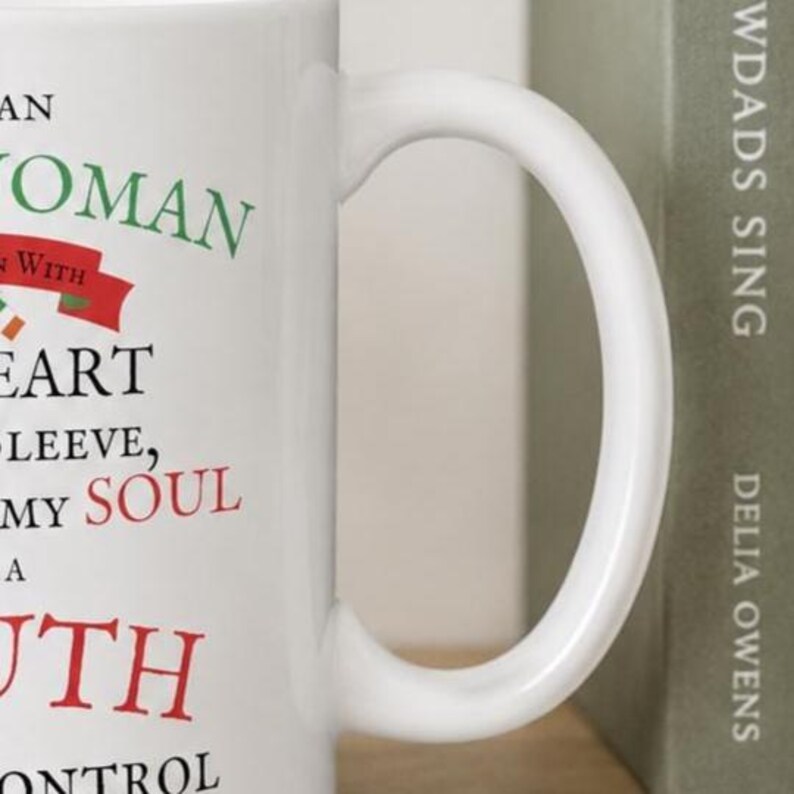 May include: A white ceramic mug featuring black, green, and red text that reads: AN WOMAN WITH HEART SLEEVE, MY SOUL A UTH ONTROL. The mug has a curved handle and a smooth finish, displaying a portion of the printed message on its side surface.