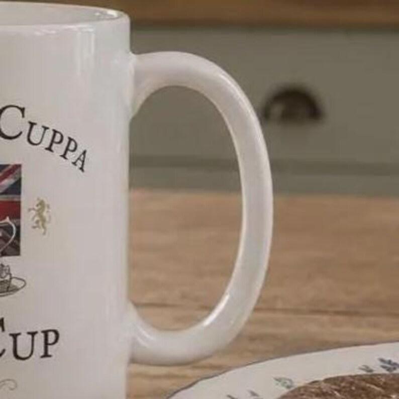 May include: A white ceramic mug featuring a curved handle and black text that reads "CUPPA" and "CUP". The design includes a small graphic of the Union Jack flag, a gold lion emblem, and a detailed illustration of a teacup and saucer on the side.