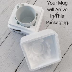 May include: A white ceramic mug nestled inside a custom-fitted white styrofoam protective box, with a separate matching styrofoam lid placed nearby. The text "Your Mug will Arrive in This Packaging." is printed in black on the right side.