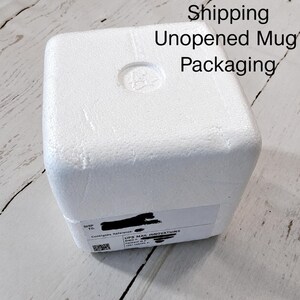 May include: A white, square polystyrene foam block used for shipping an unopened mug. The top surface has a circular indentation, and a white shipping label with black text and a QR code is adhered to the side of the protective packaging.