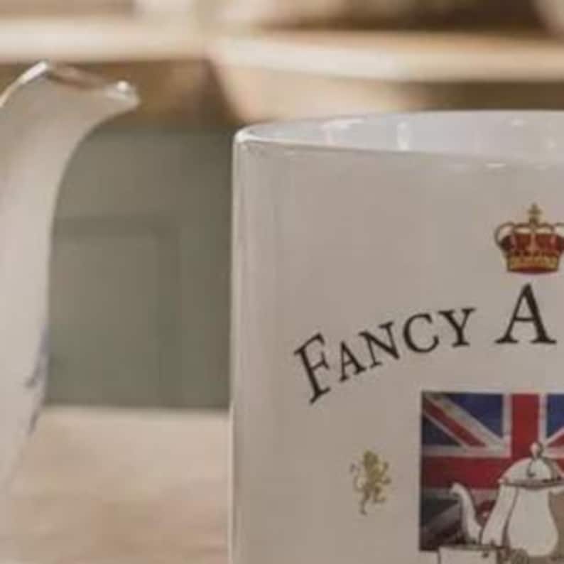 May include: A white ceramic mug featuring the text FANCY A, a red and gold crown graphic, a small gold lion emblem, and a rectangular image of a Union Jack flag with a white teapot illustration positioned below the text on the front surface.