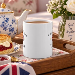 May include: A white ceramic mug featuring the text Fancy a cuppa and Tea Time, accompanied by a small Union Jack flag and gold lion icons. The mug is filled with a brown liquid and is positioned on a wooden tray next to a scone and a teapot.