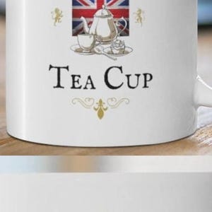 May include: A white ceramic mug featuring a graphic of a Union Jack flag, a silver teapot, a teacup, and a cupcake. The text reads "Fancy A Cuppa" above the flag and "Tea Cup" below, with a small crown icon at the top and gold flourishes.