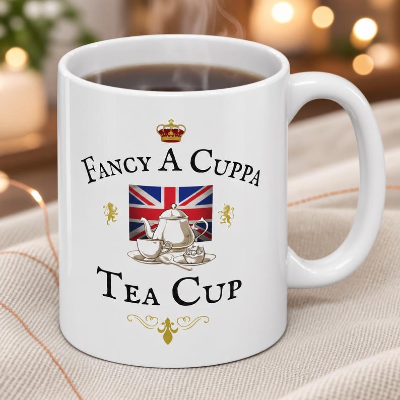 May include: A white ceramic mug featuring a graphic of a Union Jack flag, a silver teapot, a teacup, and a small pastry. The text reads FANCY A CUPPA TEA CUP, accompanied by a small crown icon at the top and a decorative gold flourish at the bottom.