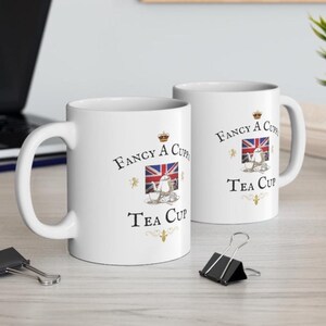 May include: Two white ceramic mugs featuring the text "Fancy A Cuppa Tea Cup" in black lettering. The design includes a small illustration of a silver teapot and a teacup centered in front of a Union Jack flag, accented with gold decorative flourishes.
