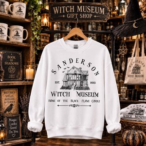 K&ouml;nnte beinhalten: Ein wei&szlig;es Langarm-Sweatshirt mit einer schwarzen Grafik eines Hauses und dem Text "SANDERSON WITCH MUSEUM EST. 1693 HOME OF THE BLACK FLAME CANDLE". Das Design zeigt eine detaillierte Illustration eines Geb&auml;udes mit einem Wasserrad.