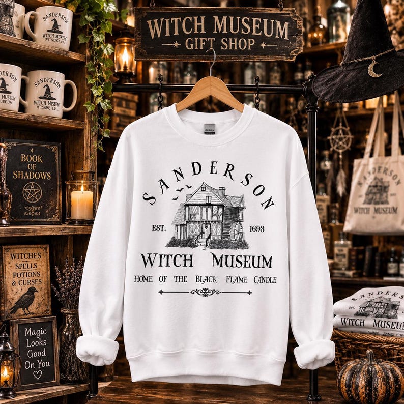 K&ouml;nnte beinhalten: Ein wei&szlig;es Langarm-Sweatshirt mit einer schwarzen Grafik eines Hauses und dem Text "SANDERSON WITCH MUSEUM EST. 1693 HOME OF THE BLACK FLAME CANDLE". Das Design zeigt eine detaillierte Illustration eines Geb&auml;udes mit einem Wasserrad.