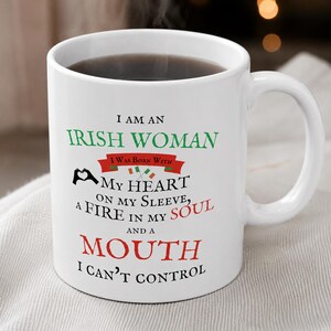 May include: A white ceramic mug featuring the black, green, and red text: I AM AN IRISH WOMAN I WAS BORN WITH MY HEART ON MY SLEEVE, A FIRE IN MY SOUL AND A MOUTH I CAN'T CONTROL, along with a small heart hand gesture and two tiny Irish flags.