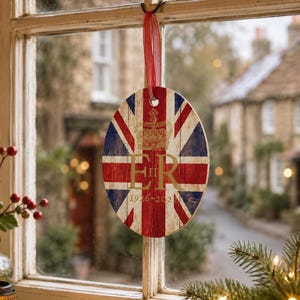 May include: An oval-shaped hanging ornament featuring a rustic wood-grain print of the Union Jack flag. The design includes a gold-colored crown graphic above the letters ER II and the years 1926-2022, suspended by a thin red ribbon.