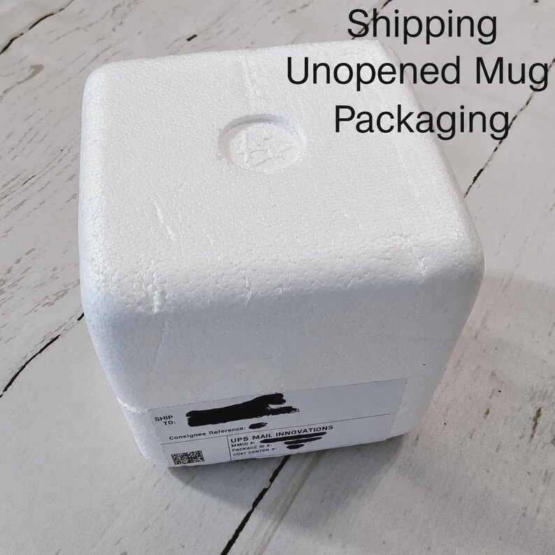 May include: A white square styrofoam shipping container for a mug. The top surface has a circular indentation in the center. A white shipping label is attached to the side with the text Shipping Unopened Mug Packaging visible above.