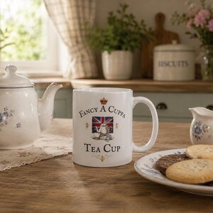 May include: A white ceramic mug featuring the text Fancy A Cuppa Tea Cup, a small crown icon, and a graphic of a teapot centered over a Union Jack flag. The design includes small gold lion accents on either side of the central teapot illustration.